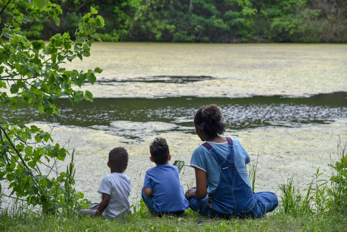 Three people are sitting on grass by a pond, facing away. One is showing something on a phone to the others. The pond is surrounded by greenery and trees, with patches of algae on the water.