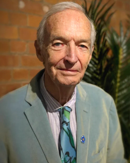 A headshot of Jon Snow wearing a light blue jacket over a striped shirt and a blue and green patterned tie with a blue Alzheimer's Society pin on his lapel. He stands against a blurred brick wall and a green plant, lit by warm light.