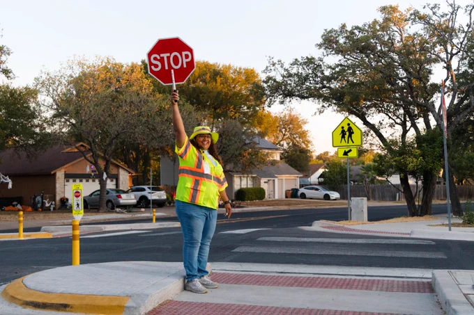 A crossing guard holding a stop sign wearing a yellow vest and hat standing in a crosswalk median.