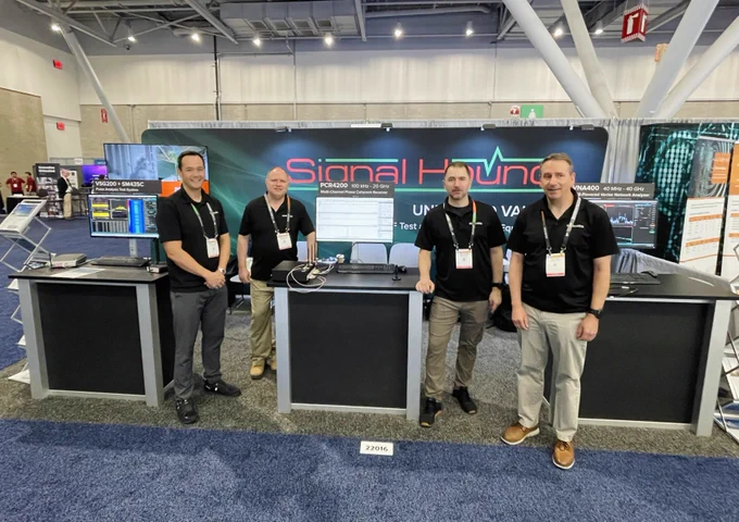 Four individuals stand at a trade show booth for Signal Hound. The booth features tech equipment and colorful displays. The setting is a large convention space with other booths visible in the background.
