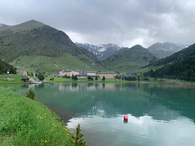 Lago de agua tranquila en primer plano con una boya roja flotando cerca de la orilla. En el fondo, un conjunto de casas, praderas verdes y montañas con parches de nieve en las cumbres. El cielo está cubierto de nubes grises y densas.