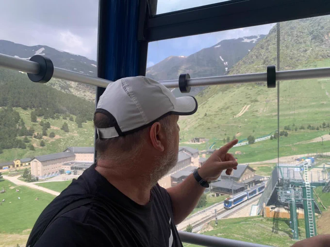 Interior de una cabina de teleférico con una persona en primer plano señalando hacia el exterior. A través del cristal se observa un entorno de montaña con praderas verdes, edificios, vías ferroviarias y una estación en la parte inferior. Las montañas del fondo presentan zonas con nieve.