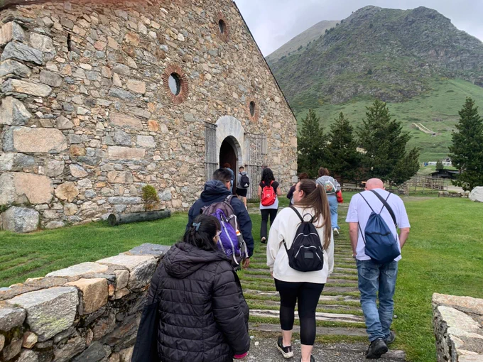 Grupo de personas caminando por un sendero de piedra hacia la entrada de un edificio de piedra con puerta arqueada. El entorno es de montaña, con césped verde, árboles dispersos y una ladera al fondo.