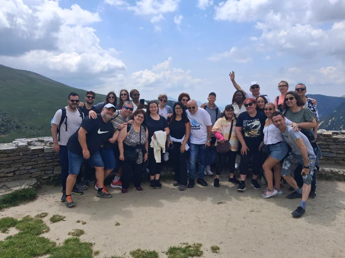 Grupo numeroso de personas posando juntas en un mirador de montaña. El grupo se sitúa delante de un muro bajo de piedra, con un paisaje montañoso y un cielo parcialmente nublado al fondo.