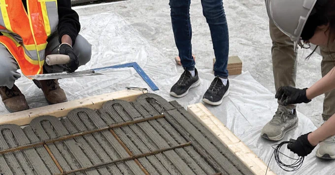 Three people are working on laying concrete over a rebar structure. One person is spreading the concrete with a trowel, another holds wiring, and a third is observing. They are standing on a plastic sheet, and one person is wearing a high-visibility vest.