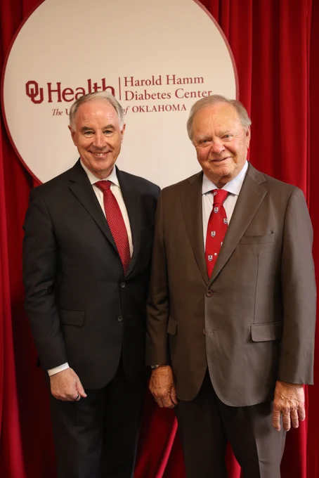 John P. Kirwan (left) with Harold Hamm in suits stand in front of a red curtain and a sign for the OU Health Harold Hamm Diabetes Center at the University of Oklahoma. Both are smiling. Photo by Abigail Gordon.