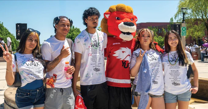 A group of individuals stands outside posing for a photo with a person in a York University lion mascot costume. They are wearing white "We Walk" T-shirts. The background features trees and a building.