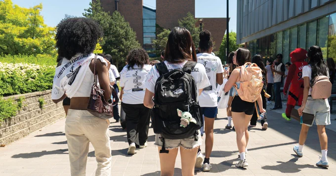 A group of people walking on a sunny path toward a brick building, many wearing matching shirts and backpacks. Some have plush toys attached to their bags. The scene suggests a casual, social setting.