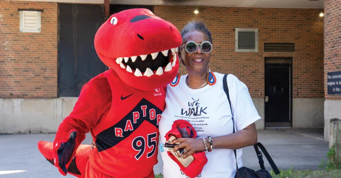 A person is standing beside the Toronto Raptors mascot, who is dressed in a red dinosaur costume with a jersey number 95. They are both smiling and posing for the photo outside a brick building. The person is holding a red item and wearing a "We Walk" T-shirt and sunglasses.