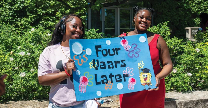Two people smiling outdoors, holding a colorful sign that reads "Four Years Later" with images of jellyfish and SpongeBob SquarePants. Lush greenery is in the background.