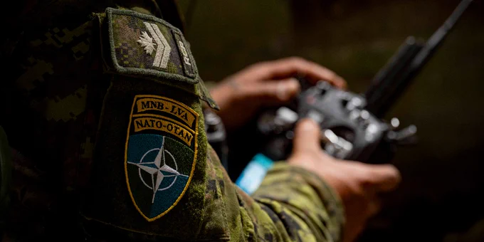 Close-up of a soldier's arm wearing a NATO-OTAN patch and holding a radio device in a dimly lit environment.