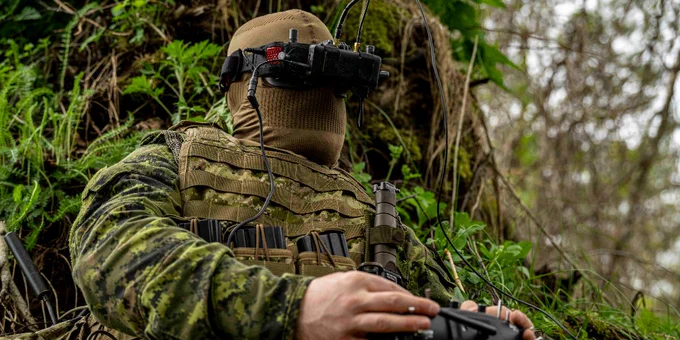 Soldier in camouflage gear operating a remote control device while wearing first person view goggles in a forest setting.
