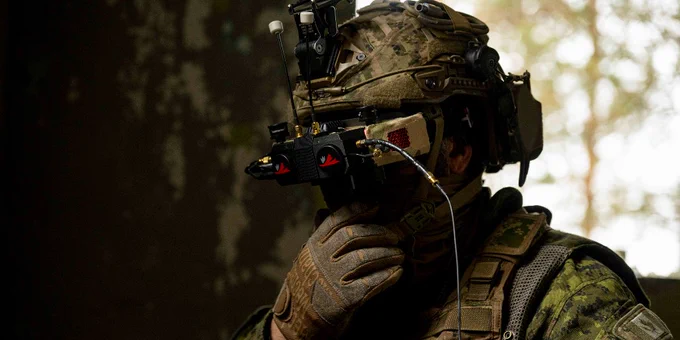 Soldier in full camouflage gear adjusting first person view goggles in a dimly lit environment.