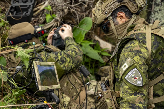 Two soldiers in camouflage gear operate electronic equipment in a dense forest setting, focusing on a device with a screen and antenna.
