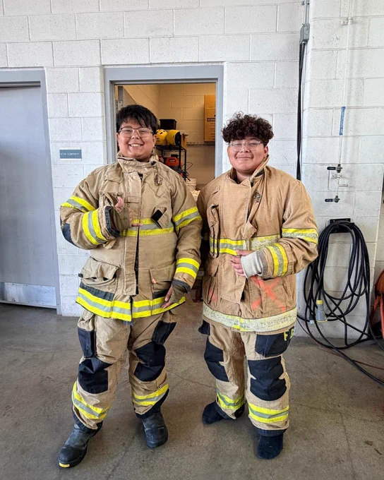 Two firefighters in protective gear standing together in a fire station.