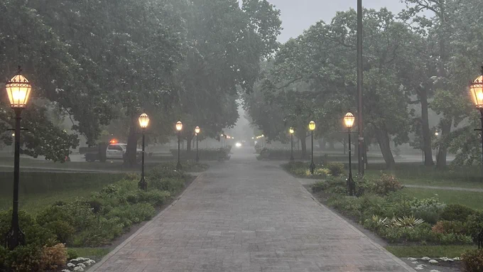 A paved pathway flanked by illuminated streetlights stretches into a rainy, tree-lined park. A white utility truck with amber flashing lights is visible in the middle ground on the left.
