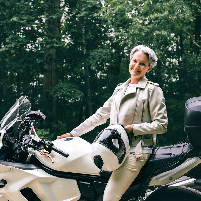 A person stands beside a white motorcycle, holding a white helmet. They are wearing a light-colored jacket, and trees can be seen in the background.