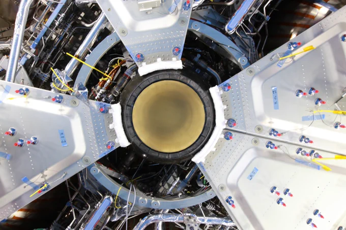 The third European Service Module's main engine being installed at Airbus in Bremen. A black circle with a golden interior; from there silver triangular segments radiate outwards with wiring that can be seen between the panels.