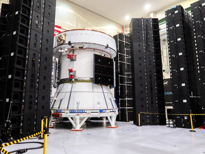 The third European Service Module and Crew Module Adapter undergoing acoustic testing at NASA's Kennedy Space Center. A large white structure - cylindrical on the top and conical on the bottom, with a black square on the cylindrical part that is a solar array - surrounded by tall black towers of speakers in a cleanroom.