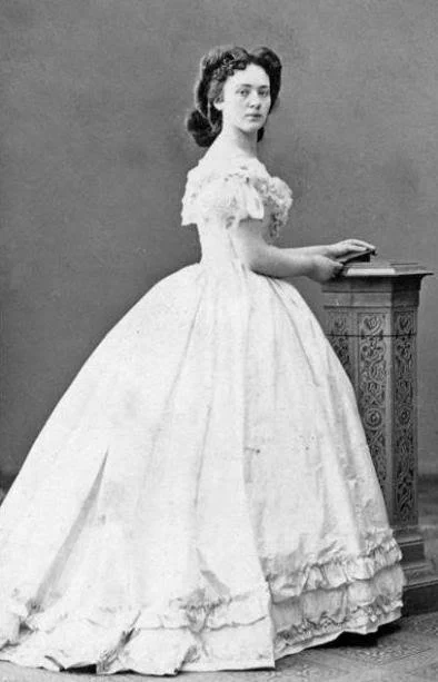 Historical photograph of Bertha von Suttner in an elegant white dress, standing beside a small decorative table.