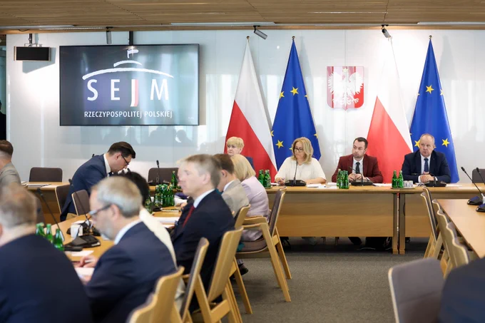 Zdjęcie sytuacyjne. Uczestnicy posiedzenia komisji obradują w sali konferencyjnej. Na pierwszym planie, z lewej strony widoczny jest fragment stołu konferencyjnego, przy którym siedzą uczestnicy posiedzenia. Na dalszym planie, przy stole prezydialnym siedzą przewodniczący i zastępcy przewodniczącego komisji. Za nimi, na ścianie, widoczne Godło Rzeczpospolitej Polskiej. Po bokach flagi Polski i Unii Europejskiej. Po lewej stronie na ścianie widoczny ekran z logo Sejmu Rzeczypospolitej Polskiej. 
