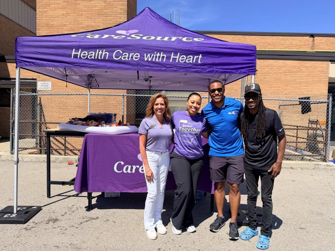 A group of four people stands in front of a CareSource booth. The booth has a purple canopy with the words "CareSource Health Care with Heart" displayed. The background includes a brick building and a chain-link fence.