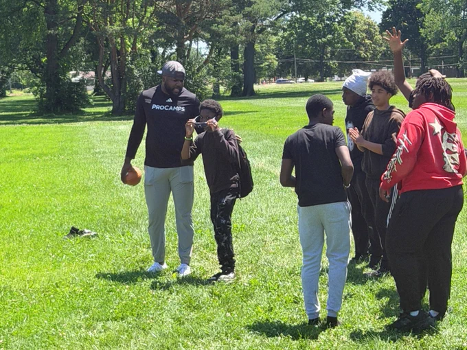 A group of people stand on a grassy field, some holding a football. One person is wearing a "ProCamps" shirt. Tall trees are visible in the background.