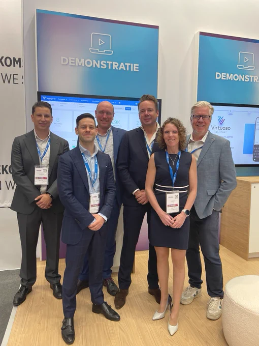 A group of six people stands smiling in front of a display booth with "DEMONSTRATIE" written on it. They are dressed in business attire and wearing conference badges. Two screens in the background show the branding for "Virtuoso.