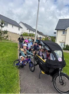 Group of schoolchildren wearing cycle helmets on bikes on a pathway. The pathway is between two grass verges and houses are in the background.