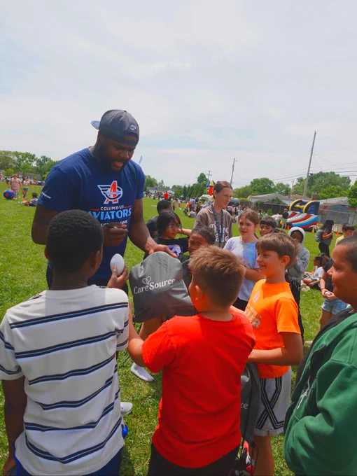 A person in a navy blue t-shirt and a cap is interacting with a group of children outside on a grassy field. They are handing out items branded with "CareSource" to the children. Several children are reaching out to receive the giveaways. In the background, more individuals are gathered on the field under a partly cloudy sky.