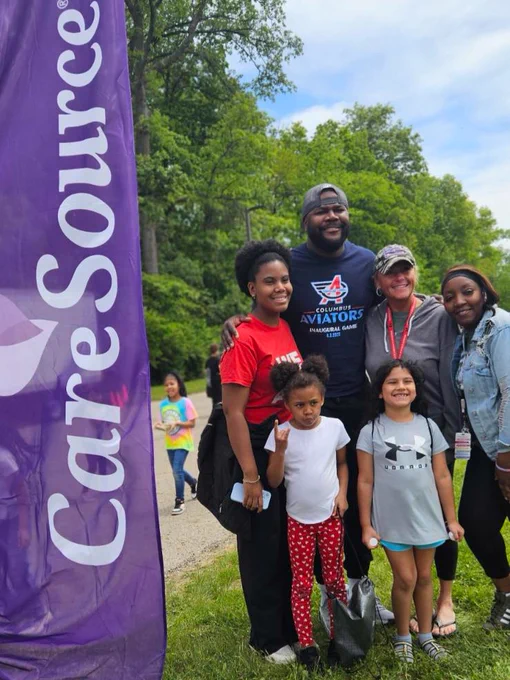 A group of six people, including four children and two adults, stand together in an outdoor setting with trees in the background. They are smiling near a vertical banner with "CareSource" written on it. Some people are dressed casually.