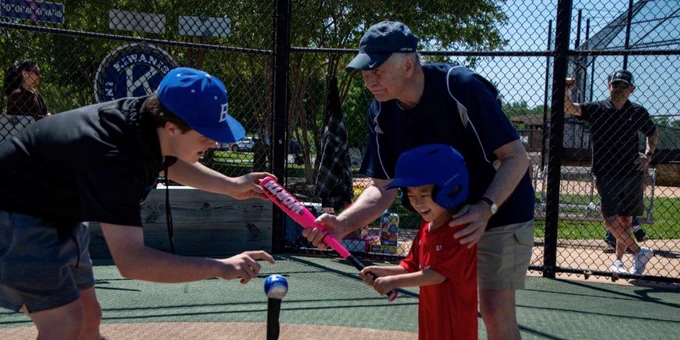 Young boy in a red shirt learning to hit a baseball off a tee with help from two adults at a sunny baseball field.