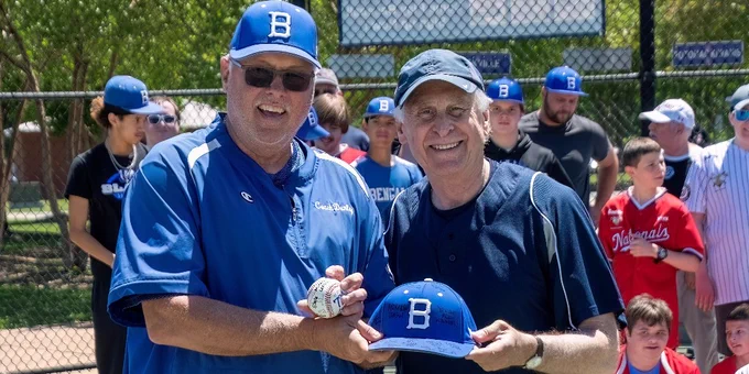 Two men in blue baseball attire holding a blue cap with a white 'B' on a sunny baseball field with a crowd behind them.