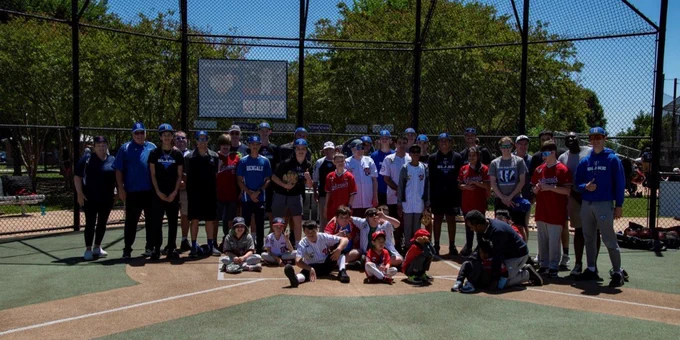Large mixed-age group gathered on a baseball diamond with a chain-link backstop and trees in the background.