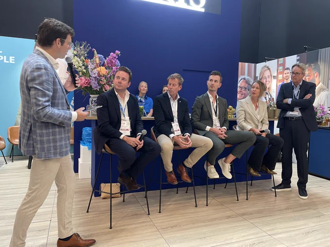 A group of individuals is seated on high stools engaged in a panel discussion. One person is standing, holding a microphone, and appears to be moderating. The backdrop features colorful floral arrangements, and there are display panels with various portraits behind them.