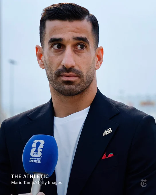 Iranian national soccer team captain Ehsan Haji Safi talks to the press while wearing a blazer and a pin that reads "#168."