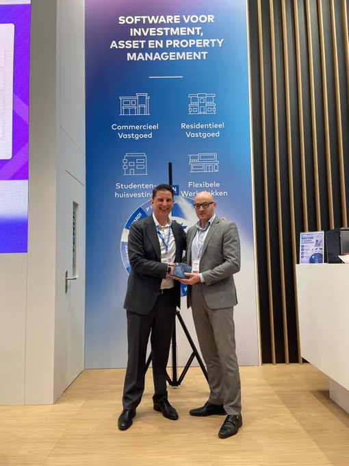 Two individuals in formal attire are standing in front of a display with a blue backdrop featuring text about software for investment and asset management in Dutch. They are both holding a plaque. A spinning wheel with various sectors mentioned is part of the display. The setting appears to be a professional event or exhibition.