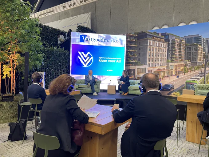 A group of people attending a panel discussion in a conference area. The backdrop features a “Vastgoedmarkt” sign and a large screen displaying a presentation titled “Are we ready for AI in real‑estate technology?” The setting includes greenery, a model or projection of urban buildings, and individuals using headphones.