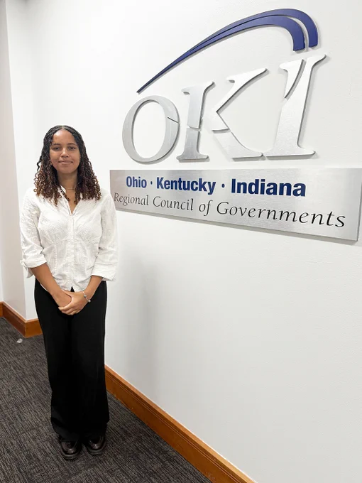 Person standing next to a wall sign for the Ohio Kentucky Indiana Regional Council of Governments in a professional setting.