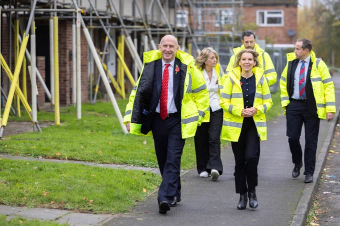 Natalie Elphicke Ross OBE with John Healey, Secretary of State for Defence, walking towards the camera on a pavement. Both wear high vis jackets as do the three people walking behind them. The houses they are walking past are covered in scaffolding. 