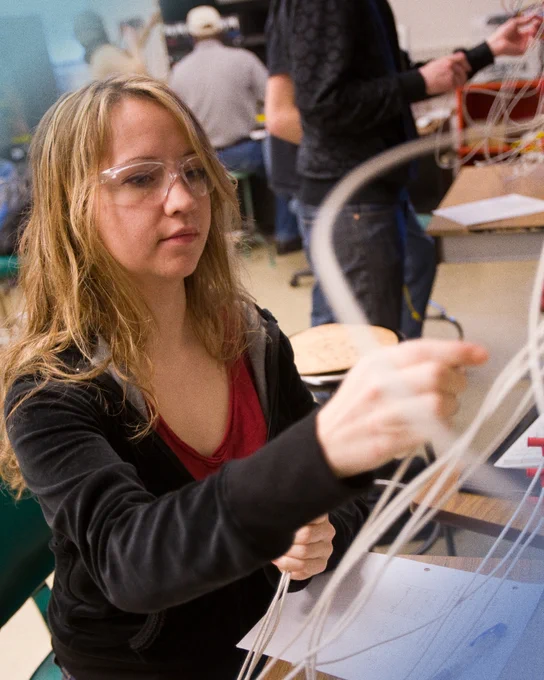 Alt text: A student wearing safety glasses carefully works with electrical wiring during an electronics lab activity, focusing on connecting components at a workstation while classmates work in the background.