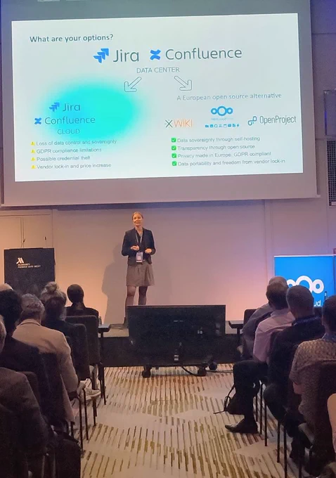 Birthe Lindenthal, Co-founder and CMO of OpenProject, speaking on stage at the Nextcloud Summit 2026 in Munich. Her presentation focuses on open source alternatives to proprietary project management and collaboration tools, with an audience attending the session.