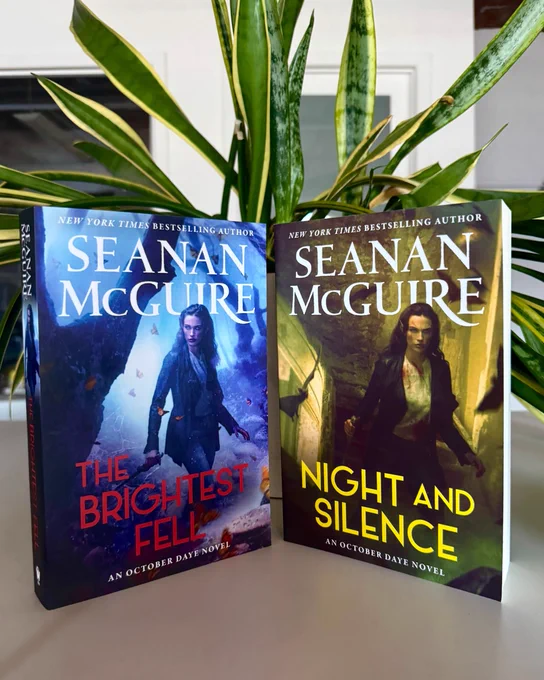 paperback of The Brightest Fell and Night and Silence on a table and in front of a plant