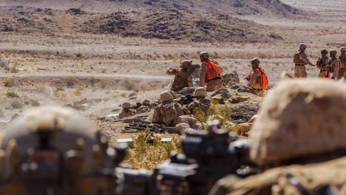 Marines establish a firing position during a live-fire training exercise in a desert environment while safety personnel observe nearby.