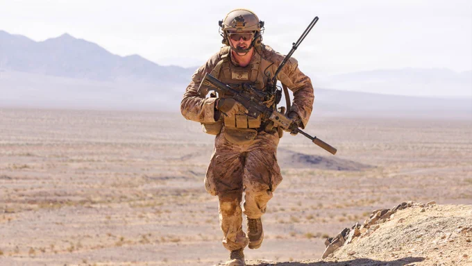A Marine runs across rocky terrain carrying a rifle during a live-fire maneuver exercise at a desert training range.