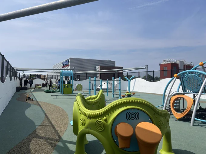 The image shows a colorful and spacious playground area on a rooftop, featuring various play structures like slides and climbing frames. In the background, the NASCAR Hall of Fame building is visible. The sky is clear with a few clouds.
