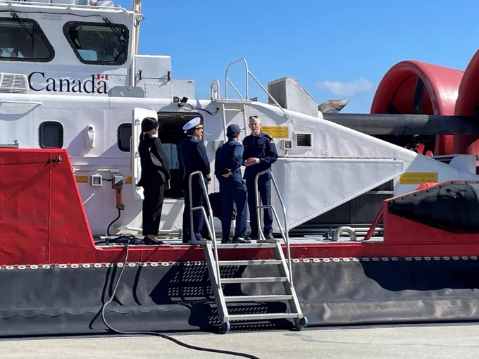 Three persons stand on the deck of a hovercraft near a metal staircase and white structures under a sunny sky.