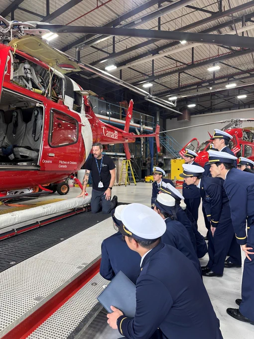 Group of uniformed members gathered in a hangar, listening to a person presenting by an open red helicopter with visible internal components.