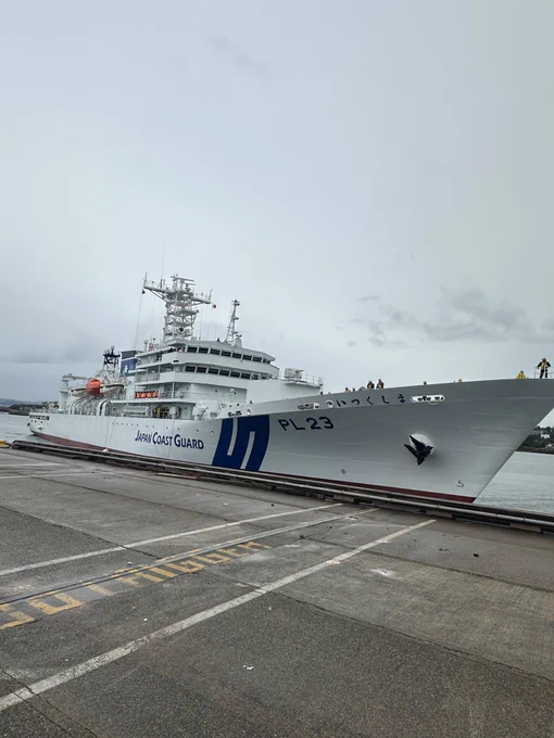 Large Japan Coast Guard white vessel docked at a concrete pier with visible mooring lines, under a grey, overcast sky. The vessel has blue markings on the hull and equipment on deck, sitting on calm water.