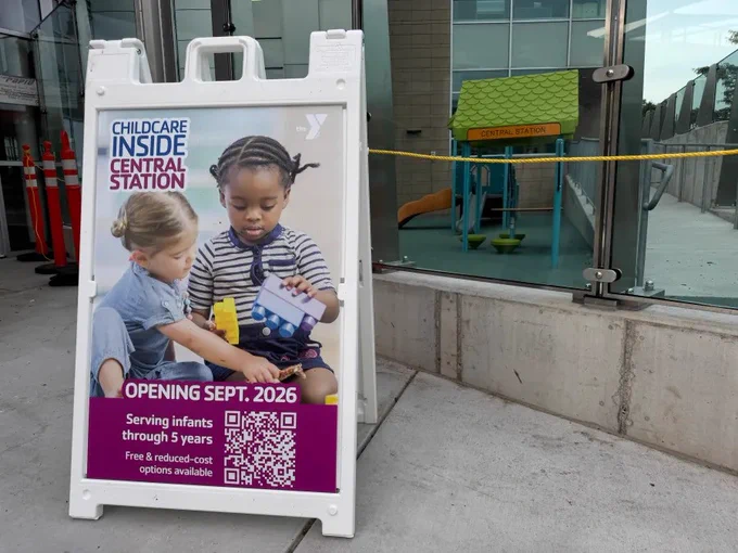This image shows a sign outside a modern building. The sign advertises "Childcare Inside Central Station" with an opening date of September 2026. It features two children playing with toys. The sign mentions services for infants through 5 years and offers free and reduced-cost options. There's a QR code on the sign, and in the background, a playground is visible.
